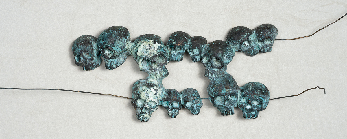 Scultura in bronzo: un filo di metallico tiene uniti dei teschi in bronzo disposti su due file.
