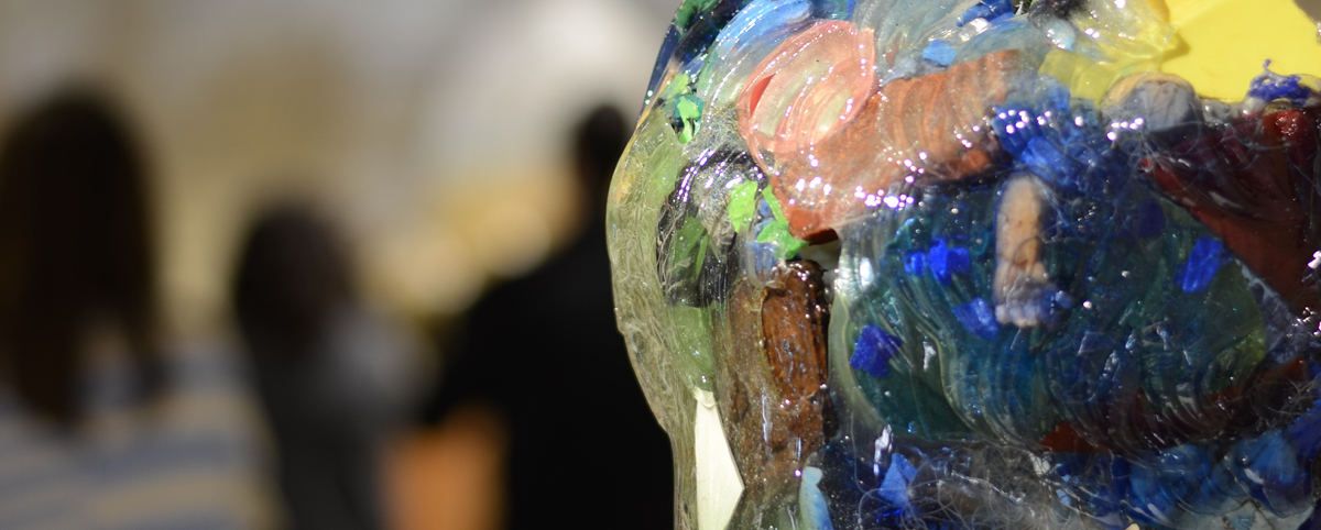 Dettaglio dell’opera Il lato oscuro del mare di Paolo Nicolai: una superficie trasparente e lucida ingloba frammenti colorati di plastica; sullo sfondo visitatori sfocati al museo.