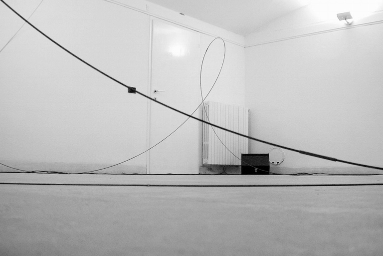 Installazione minimalista in una stanza bianca: lunghi cavi neri tesi attraversano lo spazio formando linee curve e diagonali tra pareti e pavimento. Presente un piccolo altoparlante sullo sfondo.