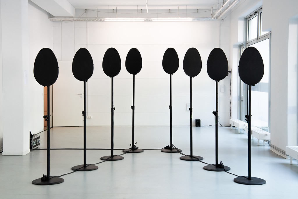Installazione É-temen-an-ki di Ramona Ponzini, opera finalista della seconda edizione del Premio Omero. Otto elementi ovali neri, sono montati su sottili aste verticali con basi circolari, disposte a semicerchio. Cavi neri collegano gli elementi sul pavimento. L’opera è allestita in uno spazio espositivo luminoso, con pareti bianche e ampie finestre sulla destra.